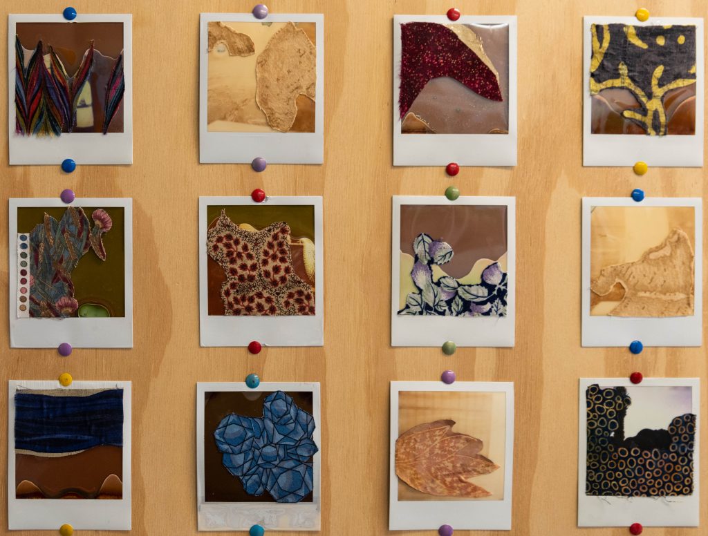 Image: Detailed view of one of the three square wood panels from 'Erode De-Compose,' zoomed into nine of the Polaroid collages. Each collage is mounted with colorful thumb tacks. The collages are all different, several with colorful patches of patterned fabric adhered to the surface. Photo by EdVetté Wilson Jones.
