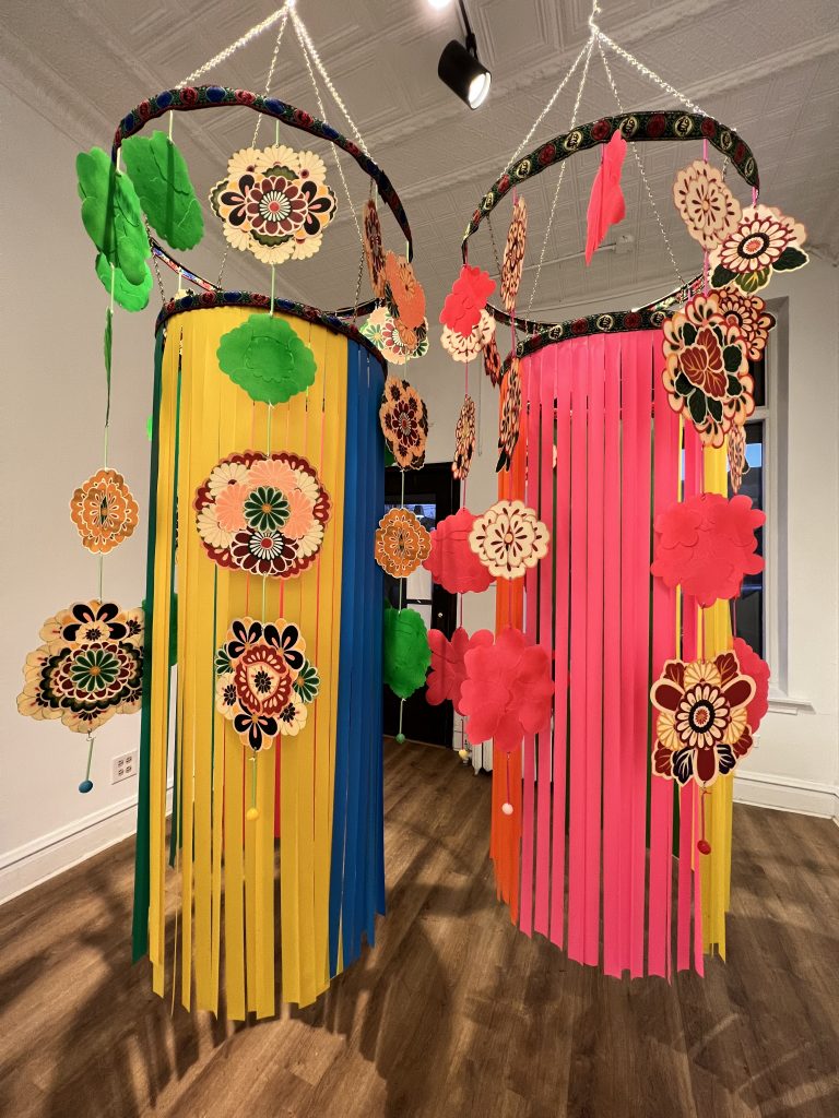 Image: Mayumi Lake, (Unison) Pink & Green Kaleidoscope, Pigment print on canvas, acrylic paint, PVC tape, metal, chain, ribbon, plastic, thread, each 84 x 24 x 24 inches, 2021. Two-embroidered hanging sculptures, with two layers hung on inner and outer circles. The outer circles has embroidered  flowers and around the smaller inner are pieces of frayed fabric.
