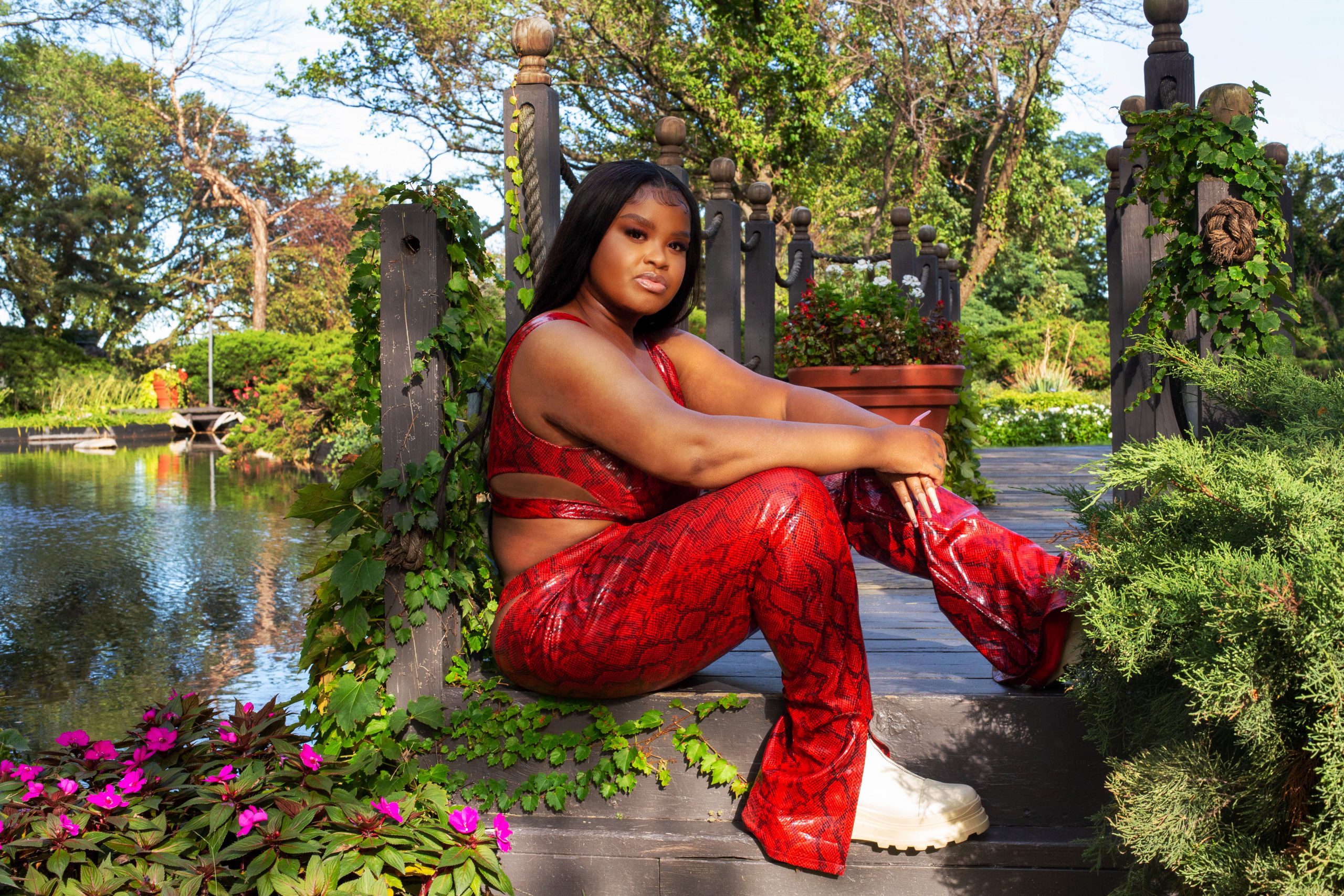 Featured image: McKensie Mack sits on a small wooden bridge in the Garden of the Phoenix or the Osaka Garden in Jackson Park, Chicago. They are wearing a bright read snakeskin ensemble with white thick-soled shoes. They have their hands resting on one leg, looking directly into the camera. Photo by Ireashia M. Bennett.