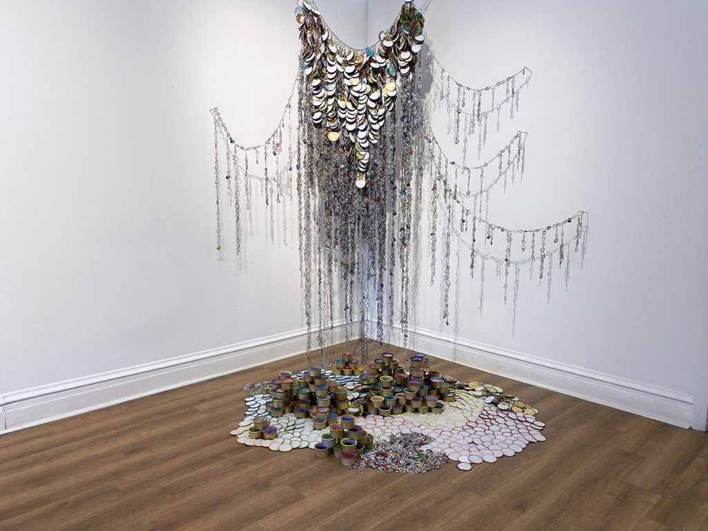 Image: Stacia Yeapanis, The Bounty and Burden of Caretaking, embroidered cat food cans and aluminum can tabs, 2021. A two-part sculpture installed both on a corner wall of the gallery and on the floor under the hanging piece of the multi-colored sculpture. Image courtesy of the artist and Annie Raab.