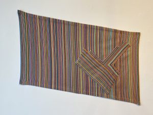 Featured Image: Stacia Yeapanis, I'm in Love with My Own Boring Life, 2022, fiber. Multicolor recetangualr fiber tapestry, hung horizontally sloped. There is a fiber motif with the same colors layered on the right side of the tapestry.