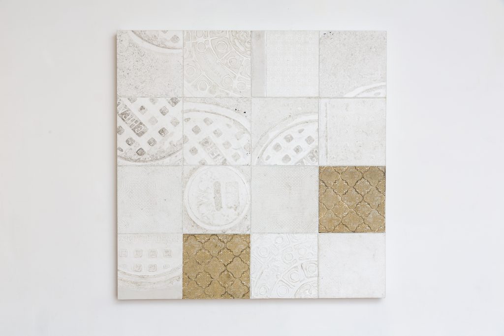 Image: "One Square Grid, 4x4" by Salvador Dominguez hangs on a white wall. There are sixteen small tiles making up one large tile. On each small tile is a relief of a manhole or object. The tiles are off-white but there are two that are a brown hue. Photo by Guanyu Xu.