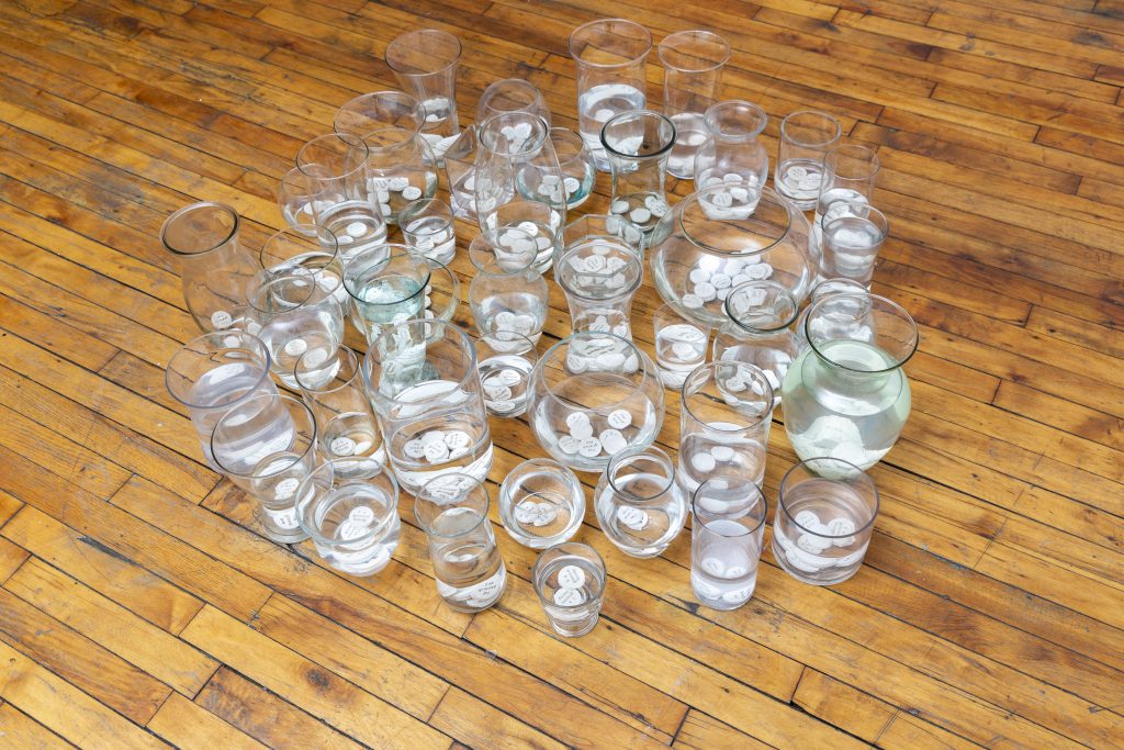 Image: Installation view of the piece "Wishing Upon a Grieving Well" by Yasmeen Nematt Alla. Clear vases and jars rest on a wooden floor with water inside of them. There are white circles —  the coins — in many of the jars. Photo by Guanyu Xu. 