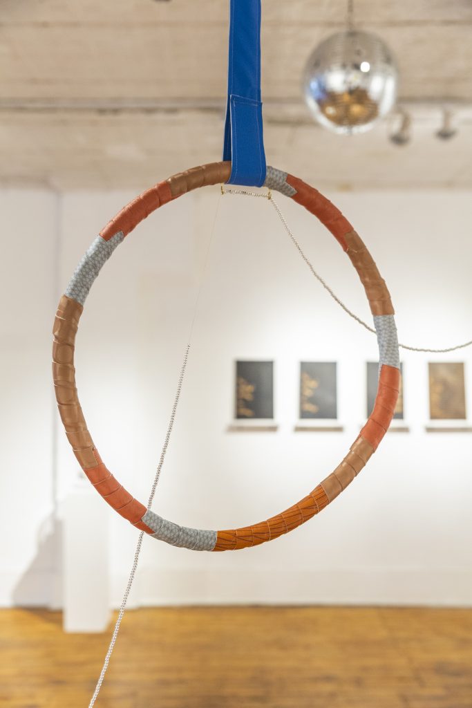 Image: "Beckon (te llamo)" by Yesenia Bell close up. A circular shape intertwined with orange and off-white thread hangs in the air. Photo by Guanyu Xu. 