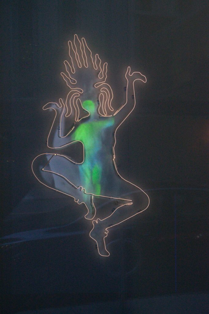 Image: Daniella Thach, As An Apsara, 2020. Krypton, transformer, projection, and artificial intelligence. The image depicts the neon outline of a dancing celestial figure from Hindu mythology. An abstract green and blue image is projection-mapped onto the negative space within the neon form. The space around the figure is dark. Photo by Nicole Thomas.