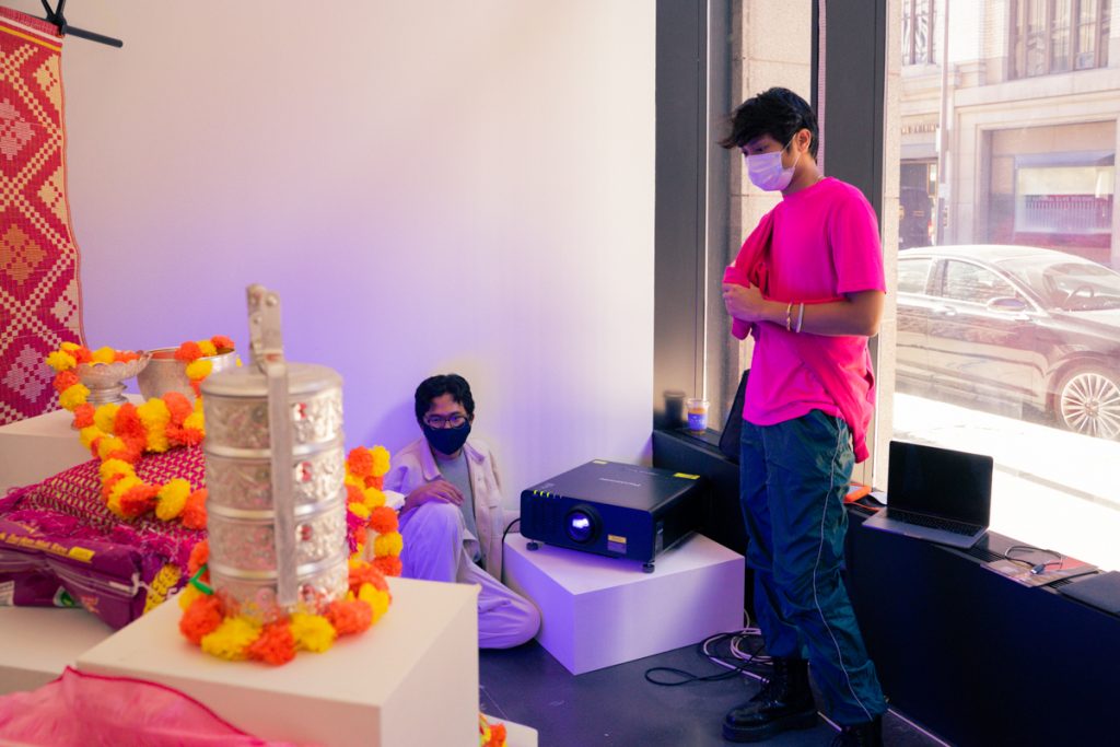 Image: Michael Khuth (right) and Sopheak Sam during installation of the piece បាត់សាសនាខ្ញុំ (Losing My Religion). Khuth, wearing a mask and a hot pink tshirt, stands next to a projector, and Sam is seated on the floor. There is a gilded gold object, a red patterned textile, bright yellow and orange garlands in the left foreground of the photo. A window behind Michael looks out onto the street of downtown St. Paul. Photo by Nicole Thomas.