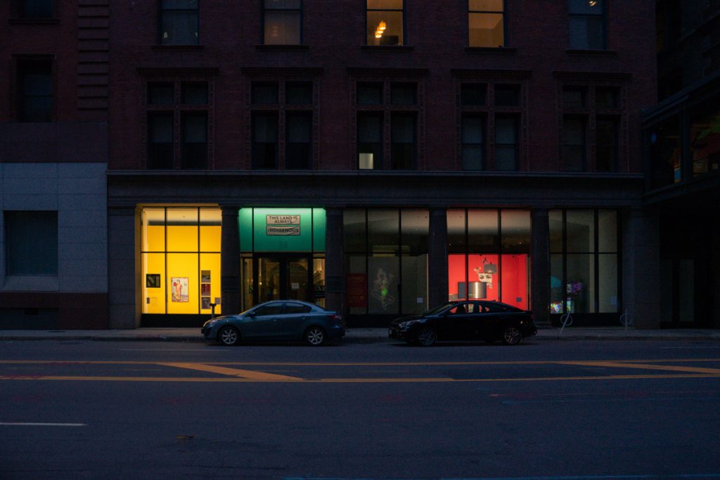 Image: A wide view of the gallery at night, a downtown facade with floor-to-ceiling windows. Cars are parked in front of the museum, foregrounding several rooms painted bright yellow, green and red. Two rooms are dark with faint colorful projections. Photo by Nicole Thomas.