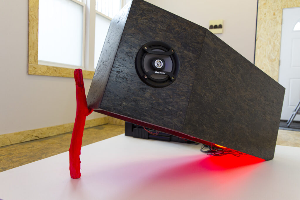 Image: Trap Coffin by Allen Moore, sound installation, OSB coffin painted black with speakers, stick painted hot pink, red LED light strip. The installation resembles a black coffin propped up with a pink stick on one side, resembling a trap. Red light comes from below the coffin. Image courtesy of Amy Shelton. 