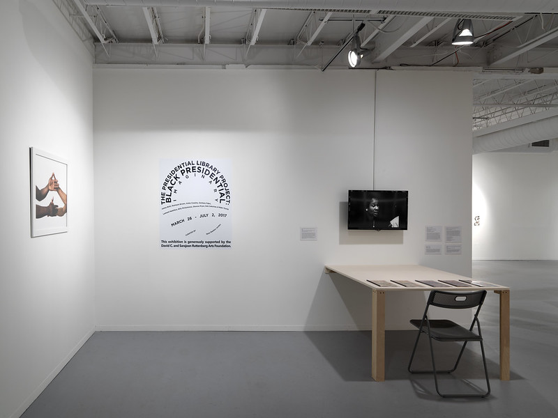 Image: Installation view of the exhibition The Presidential Library Project: Black Presidential Imaginary which was on view at Hyde Park Art Center from March 26, 2017 to July 2, 2017. To the left of the image is a photograph of two sets of hands in mid-dap, hanging on a white wall. On the adjacent wall is the title text for the exhibition shaped into the Obama presidential campaign logo, which is shown next to a monitor with a black-and-white image on the screen, just above a table and chair with booklets on the surface. Photo courtesy of Hyde Park Art Center.