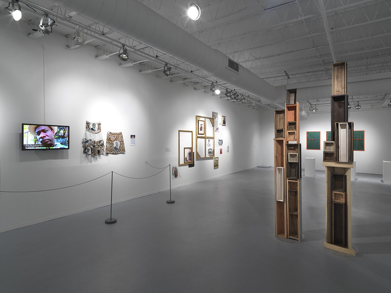 Image: Installation view of the exhibition The Presidential Library Project: Black Presidential Imaginary which was on view at Hyde Park Art Center from March 26, 2017 to July 2, 2017. To the left of the image is a monitor with a still, close-up image of a person looking directly into the camera. Next to the monitor are several wall-mounted sculptural artworks with photographic images dispersed throughout. On the adjacent wall is a series of green images framed in bright orange with bright orange, illegible text in the middle. In the foreground there are two monolithic wooden assemblage sculptures. Photo courtesy of Hyde Park Art Center.