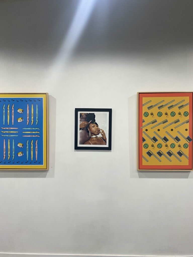 Image: An installation view of three pieces hanging on a wall as part of the show FEMME at FLXST Contemporary. The middle piece is Maiwenn Raoult's photograph The January Twins, 2021, 12 x 16". The two pieces on either side are colorful digital prints on linen by Merryn Omotayo Alakaand Sam Fresquez. The right piece is titled Oh Snap and the left is titled It's God Given. Image courtesy of FLXST Contemporary.