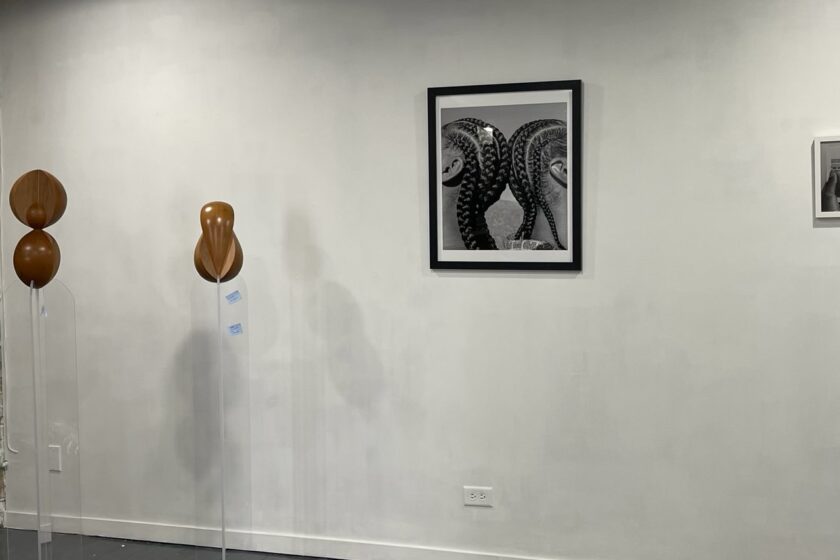 Featured image: An installation view of FEMME at FLXST Contemporary. On the left are two wooden, organic-shaped sculptures by Vivian Chiu, and on the right is a black and white photograph by Maiwenn Raoult of two people with braids standing back to back. Image courtesy of FLXST Contemporary.
