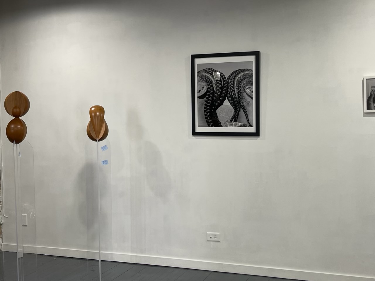 Featured image: An installation view of FEMME at FLXST Contemporary. On the left are two wooden, organic-shaped sculptures by Vivian Chiu, and on the right is a black and white photograph by Maiwenn Raoult of two people with braids standing back to back. Image courtesy of FLXST Contemporary.