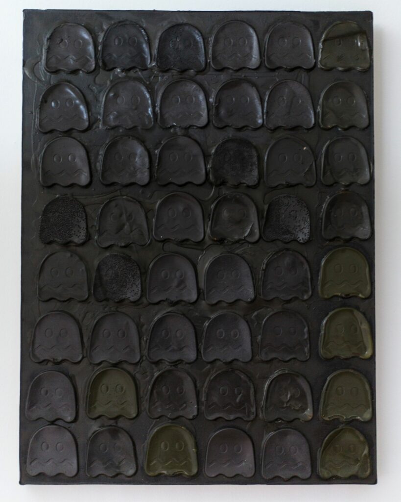 Image: Blue Shift 48 by Allen Moore, casted graphite and glue on canvas. On a white wall hangs one rectangular panel with 48 graphite and glue cast Pac-Man ghosts. Image courtesy of Amy Shelton. 