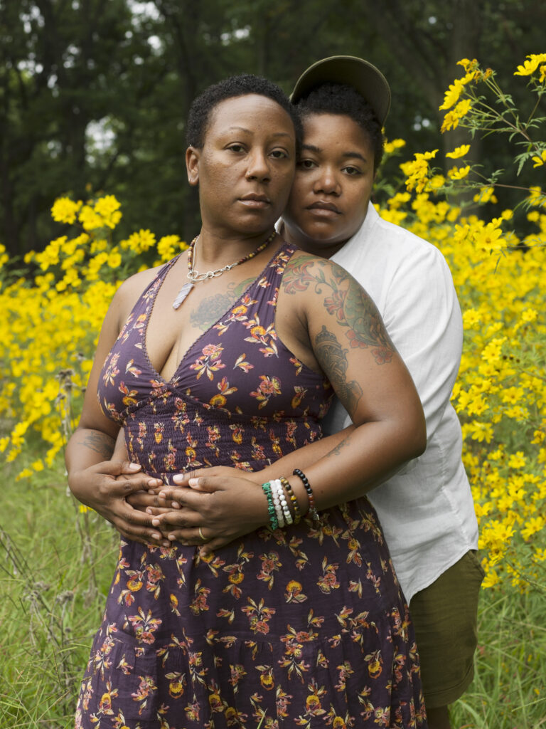 Image: Kelli and Jen, 2017. Photo by Jess T. Dugan. Set with prairie grass, yellow flowers, and some trees, two Black people in embrace, one wrapping their arms around the other and the embraced places their hands on the embracers hands, both looking at the camera. The embraced has bracelets on their left and tattoos on their left; they wear a purple dress, with a floral pattern. The embracer wears a white shirt, olive shorts, and a baseball cap. Image courtesy of the artist.