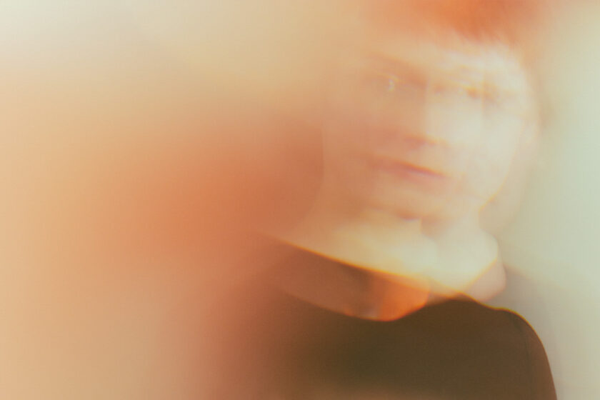 Featured image: A portrait of a person with short hair who appears to be in mid motion. Their face is slightly blurred and there is an orange-yellow haze around them. Photo by Sarah Joyce.