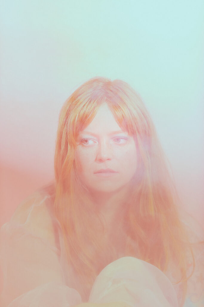 Image: Portrait of Miranda, 8/12/2021. A person with long, orange hair looks down and to the left. The hazy lighting is orange and light blue. Photo by Sarah Joyce.