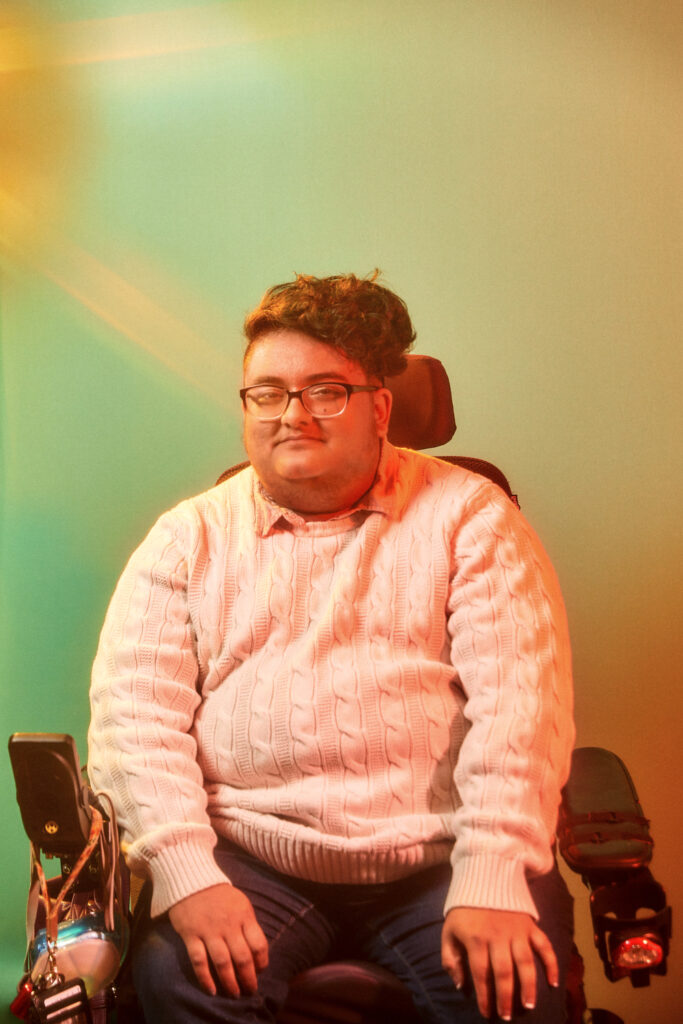 Image: Portrait of Noor Pervez, 02/19/2022. A person wearing a light sweater, glasses, and jeans sitting in a wheelchair. The background shows green and orange light. Photo by Sarah Joyce.