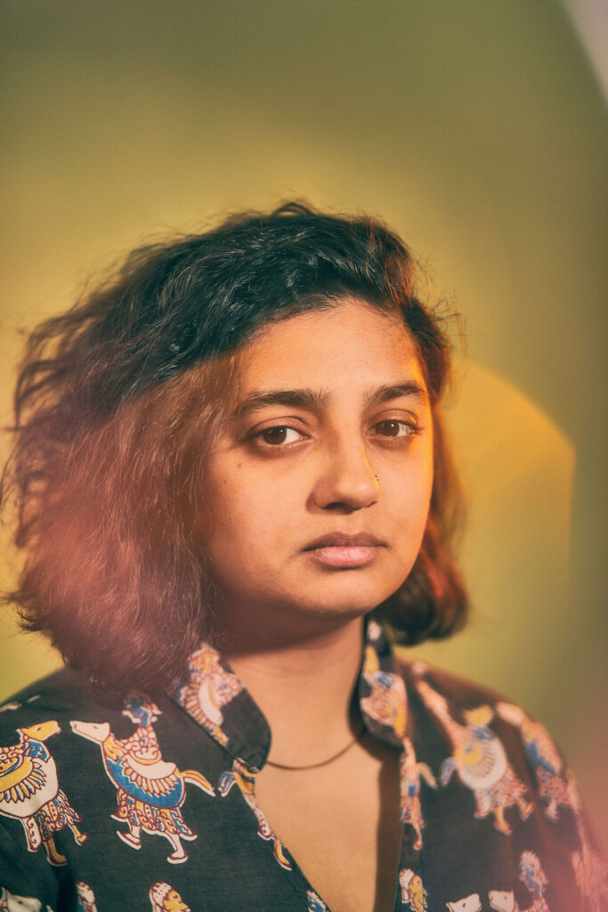 Image: Parita, 11/08/2021. A person with black hair, a nose ring, a gold necklace, and a collar shirt looks directly at the viewer. The background is largely green. Photo by Sarah Joyce.