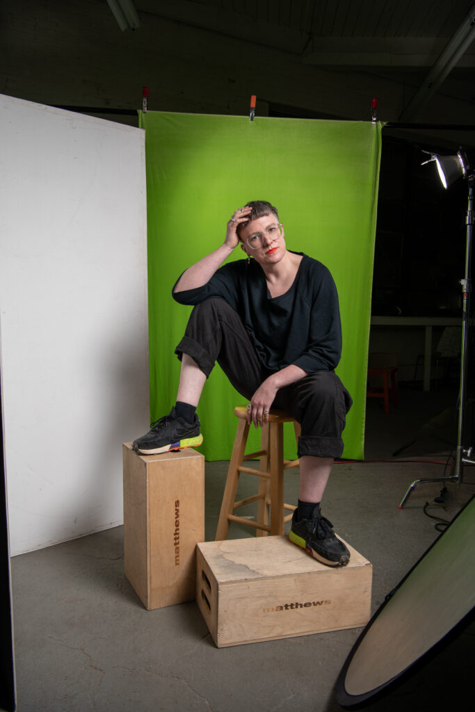 Image: Portrait of Sarah Joyce. Sarah is sitting on a wooden stool with her head in her hand. She has short hair, glasses, and red lipstick. There is a green backdrop behind her. Photo by Mariah Karson.