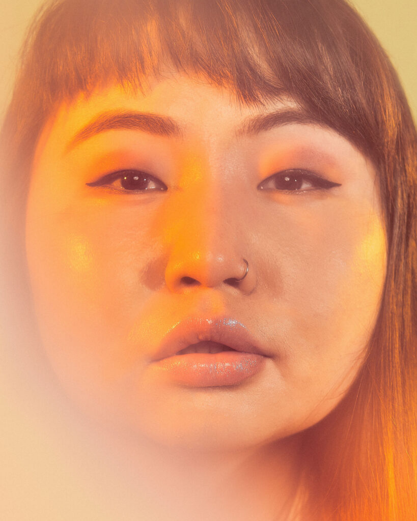Image: Portrait of Tuul, 8/21/2021. A person with long hair, a nose ring, and eyeliner looks straight at the viewer. Orange lighting shines on her face. Photo by Sarah Joyce.