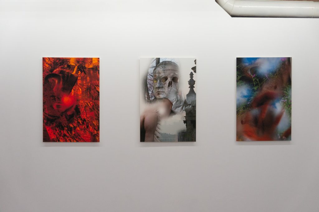 Image: Gallery wall view of Kristoffer Ala-Ketola's Dissonance, Morphing, Dreaming, 2022, pigment prints on paper, dimensions: 20” x 30”. From the left: a pigment print with mostly red and black coloring; some orange the texture of the painting resembles a forest fire; smoke and debris, which the subject is experiencing emotionally. In the second pigment print there is the same subject looking at the audience, a skull, a window with woods and a tall grave headstone. the colors are mostly shades of grey, with flesh tones. The third pigment print is the most difficult to pick out the subject; it's main colors are red, white, blue and green. There are vines with flowers and a ghostly figure falling into mist or a portal in the earth. Image courtesy of Livy Snyder.