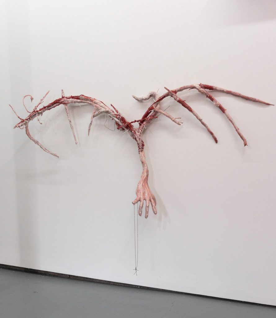 Image: Gallery view of Kristoffer Ala-Ketola's Geryon’s Wings, 2022, sculpture made out of pages of a book, wood, metal, papier-mâché, modeling clay, pigment, plaster, zip ties, and silicone. An gallery wall view  of Wings; the wings themselves are sparse and look like flesh and blood. An arm with a hands is at the center reaching towards, when the spine of the wings would be. A wire necklace with a small wired body is balanced on the pinky of the hand. Image courtesy of Livy Synder.