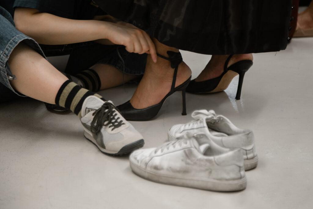Image: A close up shot of three pairs of shoes and a hand. A person is shown tying black, high-heeled shoes onto another person. A pair of white sneakers lay in the foreground, while the person on the left is wearing black and white sneakers. Photo by Abigail Teodori.