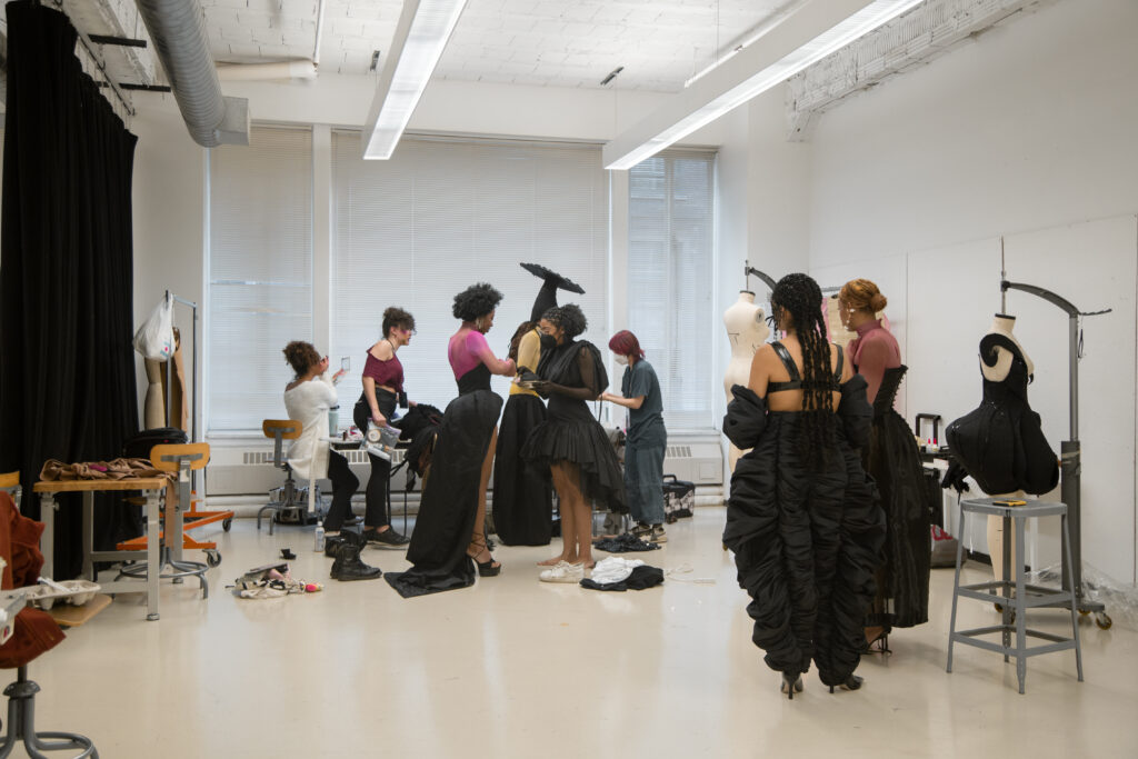 Image: A room full of people, garments, and supplies getting ready to wear and show the garments in the Iyomi Ho Ken's collection. Photo by Abigail Teodori.