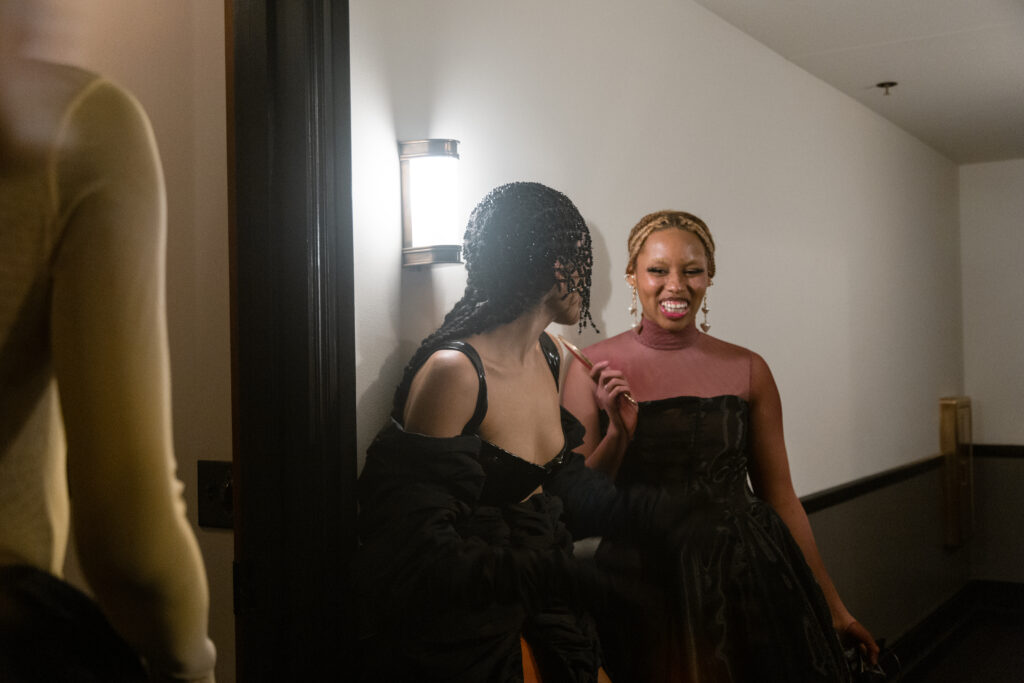 Image: Two models adorned in pieces from Iyomi Ho Ken's collection stand in a hallways be a wall light. They look at each other and smile. The model in the middle of the frame is wearing a black outfit and headpiece, while the model on the right wears a black dress with a shear pink top section. Photo by Abigail Teodori.