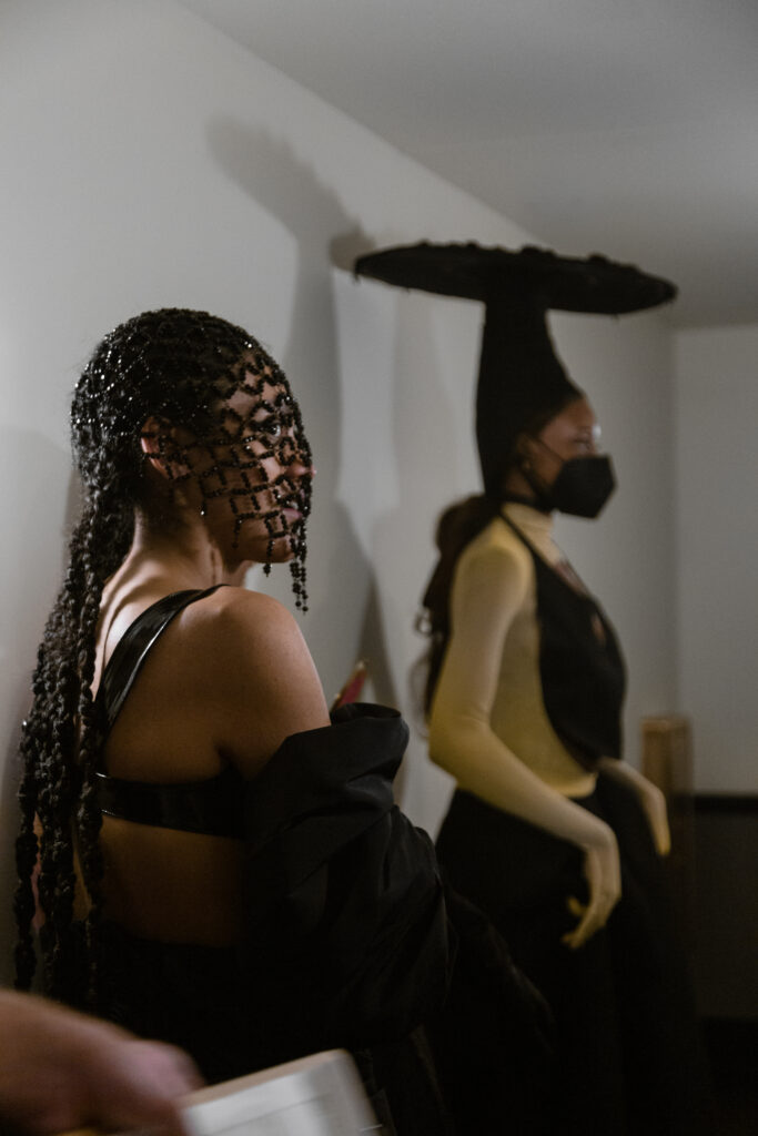 Image: Two models wear pieces from Iyomi Ho Ken's collection. The model in the foreground wears an all black outfit and a black headpiece. The model in the background wears a tall, black headpiece and black and yellow garments. Photo by Abigail Teodori.