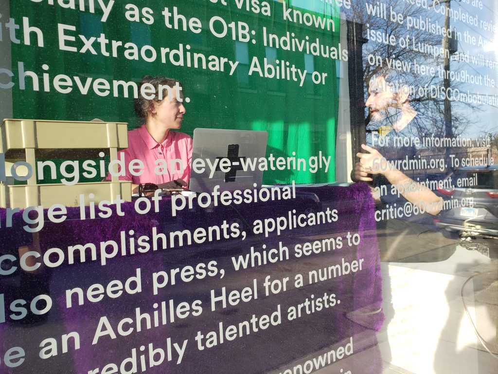 Image: Lori Waxman is seated at a desk behind a shop window. She sits in front of an open laptop as she talks to a beard person off to her left. She is slightly obscured by vinyl words printed on the shop window. 60 wrd/min performance at Co-Pro (Lori reviewing Li-Ming Hu). Photo by Li-Ming Hu.  