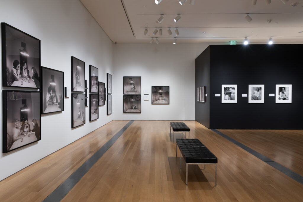 Image: Installation view of Dawoud Bey & Carrie Mae Weems: In Dialogue at the Grand Rapids Art Museum. The walls in the back, right corner of the image are black and showcase various black and whtie photographs. The walls in the back and to the left are white and showcase black and white photographs from Carrie Mae Weems' The Kitchen Table Series. Courtesy of the museum.