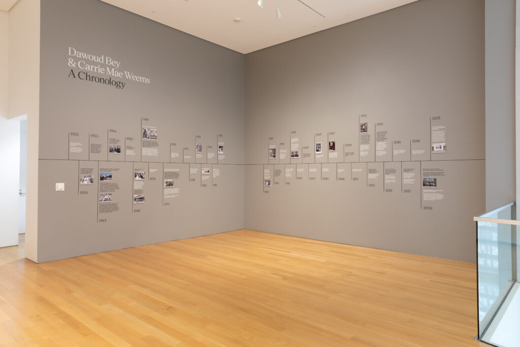 Image: Installation view of Dawoud Bey & Carrie Mae Weems: In Dialogue at the Grand Rapids Art Museum. The image shows a timeline of the two artists' lives installed on a light grey wall. Text on the top left corner reads, "Dawoud Bey & Carrie Mae Weems A Chronology". Courtesy of the museum.