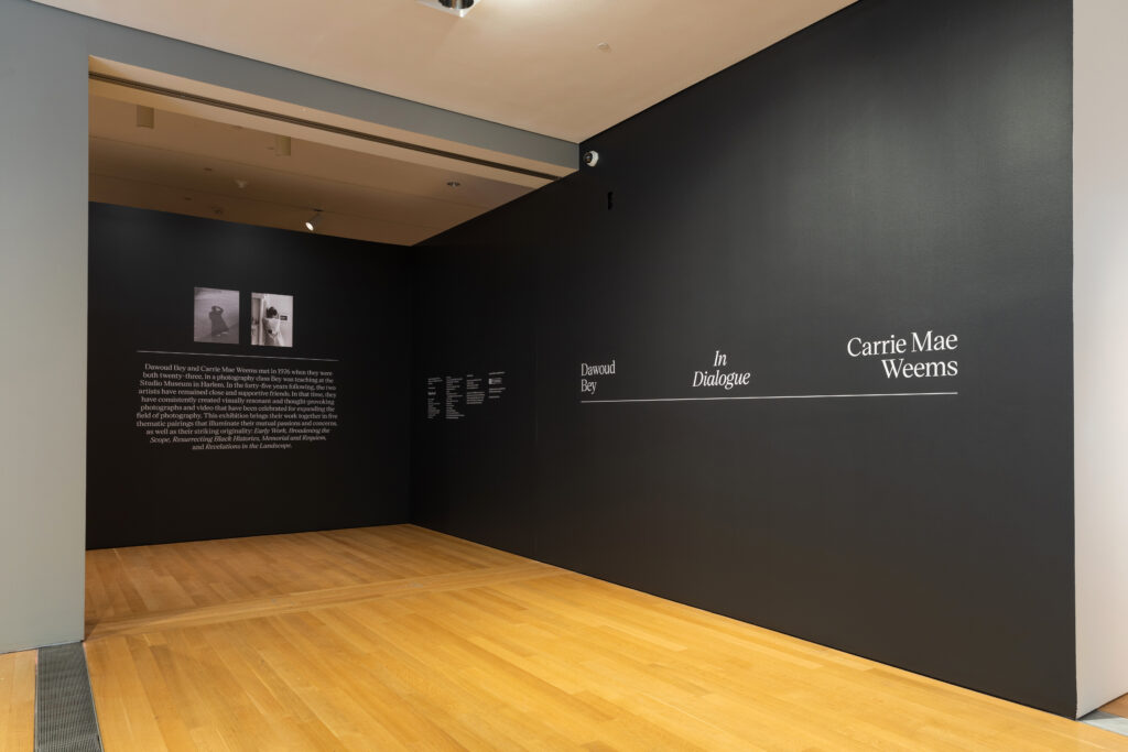 Image: Installation view of Dawoud Bey & Carrie Mae Weems: In Dialogue at the Grand Rapids Art Museum. The photo shows a black wall with white text and two black and white photographs on the back section of the wall, and text that says the title of the exhibition on the front, right section of the wall. Courtesy of the museum.