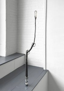 Image: G.LaPointeJr, ( western smoke ),2022, interlocking hitch ball, graphite treated spent firework cylinder and tree branch, nickel-plated steel O- rings, found empty lighter and half- cut hardened steel padlock shackle, 11 x 2 x 45.5”. A branch with bolts at the bottom standing straight up from the gallery floor with steel O - rings hangs in branch knots. A white lighter is at the top. Image courtesy of Roman Susan.