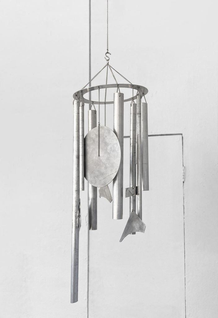 Image: Gary LaPointe Jr., ( wind chime ), 2022, repurposed aluminum, various aluminum pipes, found cut steel chain link, hardware, 10 x 12 x 25.25”. A silver metal wind chime hangs from the gallery ceiling. Image courtesy of Roman Susan. 