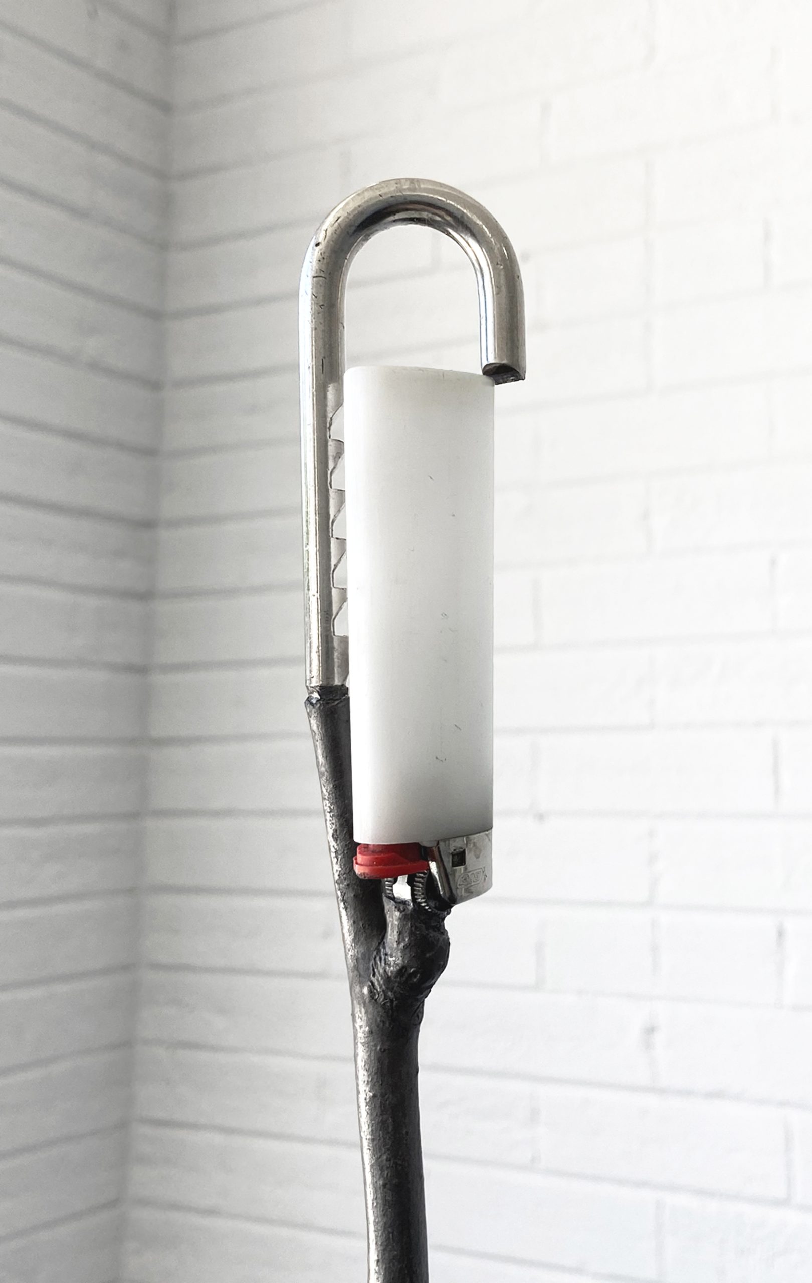 Image: Gary LaPointe Jr., ( western smoke ),2022, interlocking hitch ball, graphite treated spent firework cylinder and tree branch, nickel-plated steel O- rings, found empty lighter and half- cut hardened steel padlock shackle, 11 x 2 x 45.5”. A detail view of the top of the sculpture where a white lighter is held in the grip of a metal part attached to the branch. Image courtesy of Roman Susan.