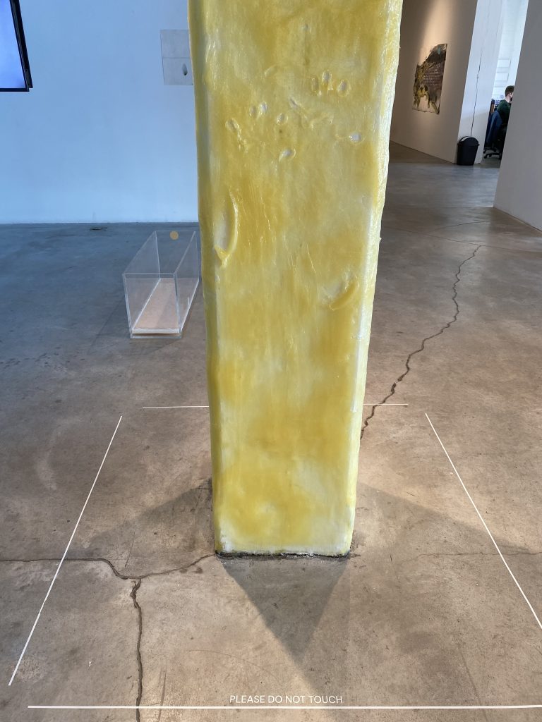 Image: Jean-Charles de Quillacq, Armpit, 2021, petroleum jelly and human armpit hair on column, dimensions vary. A white column is slathered in amber-colored petroleum jelly. Two handprint and arm prints can be seen in the jelly. The concrete floor has a white line around the artwork with the words, "PLEASE DO NOT TOUCH".