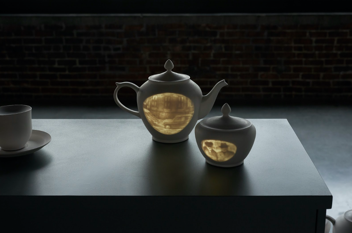 Installation shot of In Fugue by Rae Stern shot depicting a white tea kettle with an illuminated street scene and sugar pot with two men in winter clothes.