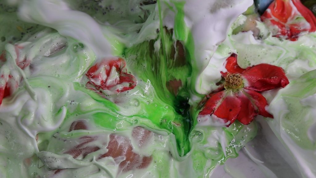 Image: Adham Faramawy, Skin Flick (still), video, 13:30 minutes. Courtesy of the artist and Bemis Center for Contemporary Arts.  Lime green and white, filmy liquid swirls around the composition. Pink flowers are saturated in the liquid.