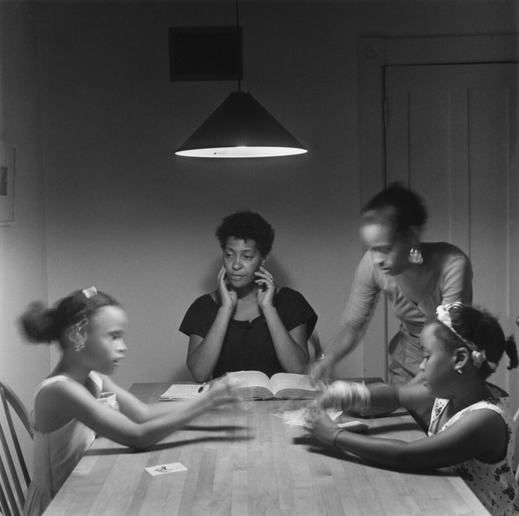 Image: Untitled (Woman and Daughter with Children) from The Kitchen Table Series, 1990. Gelatin silver print, 27 ¼ x 27 1/4. © Carrie Mae Weems. Courtesy of the artist and Jack Shainman Gallery, New York. The image shows a Black family sitting at a kitchen table. The children are playing a card game and the woman at the head of the table has a book next to her.