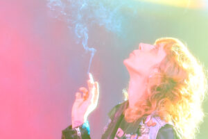 Image: A person with brightly lit curly hair in a leather jacket and denim vest smoking a cigarette, blowing smoke upwards. the background is a gradient of pink to green.