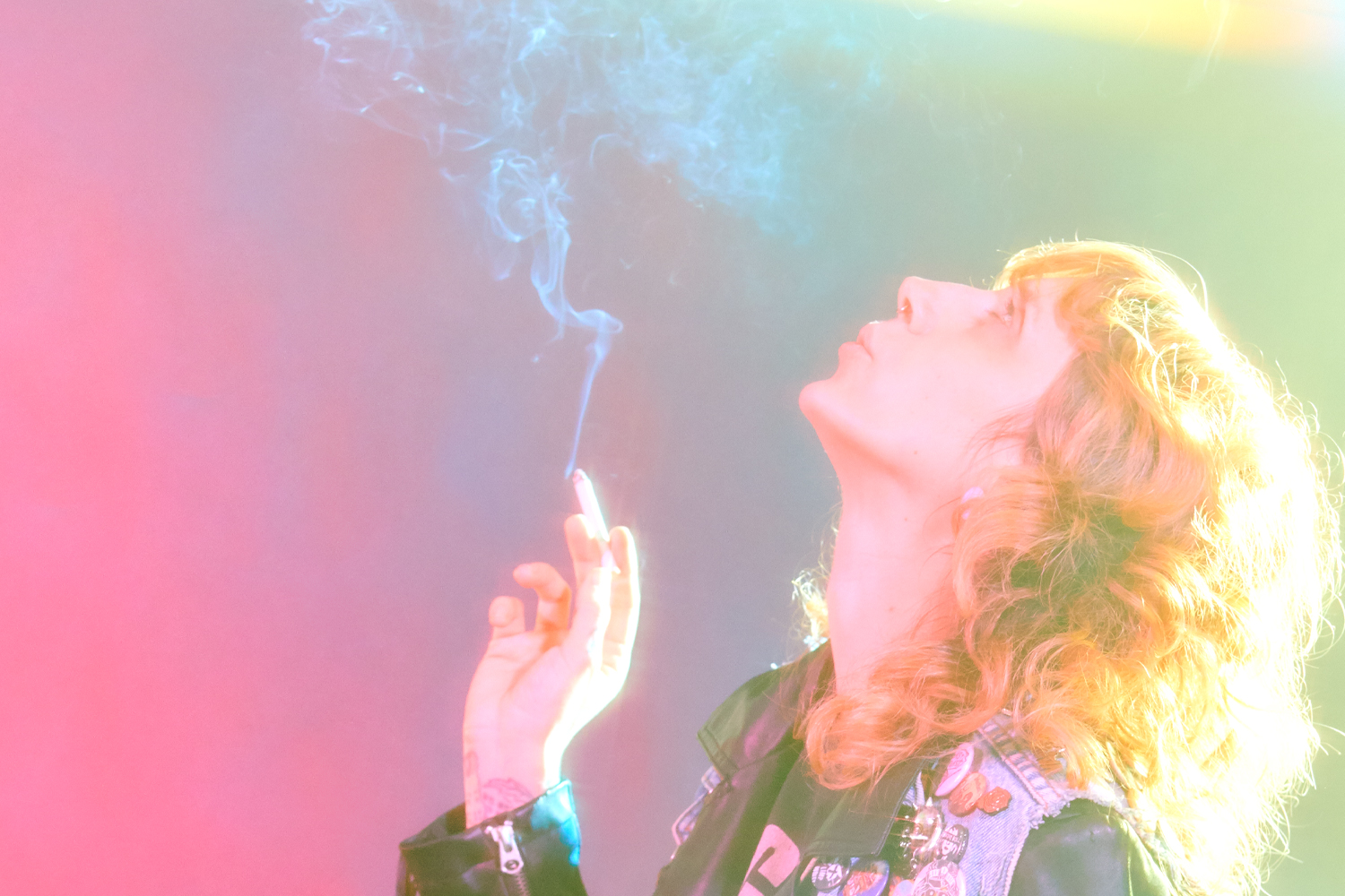 Image: A person with brightly lit curly hair in a leather jacket and denim vest smoking a cigarette, blowing smoke upwards. the background is a gradient of pink to green.