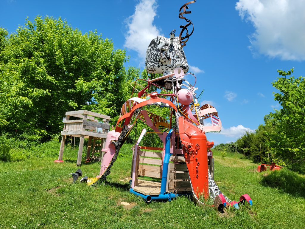 Image: A photo of a multi-colored, intricate, large sculpture with winding bits of metal at the top. A wooden structure is to the left of the sculpture. Both are surrounded by lush green vegetation. Photo by the author.