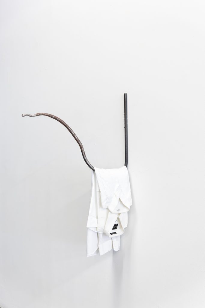 Image: Amina Ross, Rest, (Refrain), 2021 at The Luminary. Two white shirts, which also resemble white hotel towels, hang on a metal wire sculpture. The sculpture almost makes the shape of a 'U', but the end section of the metal sculpture juts out. This specific part looks worn and rusty. Image courtesy of Kalaija Mallery.
