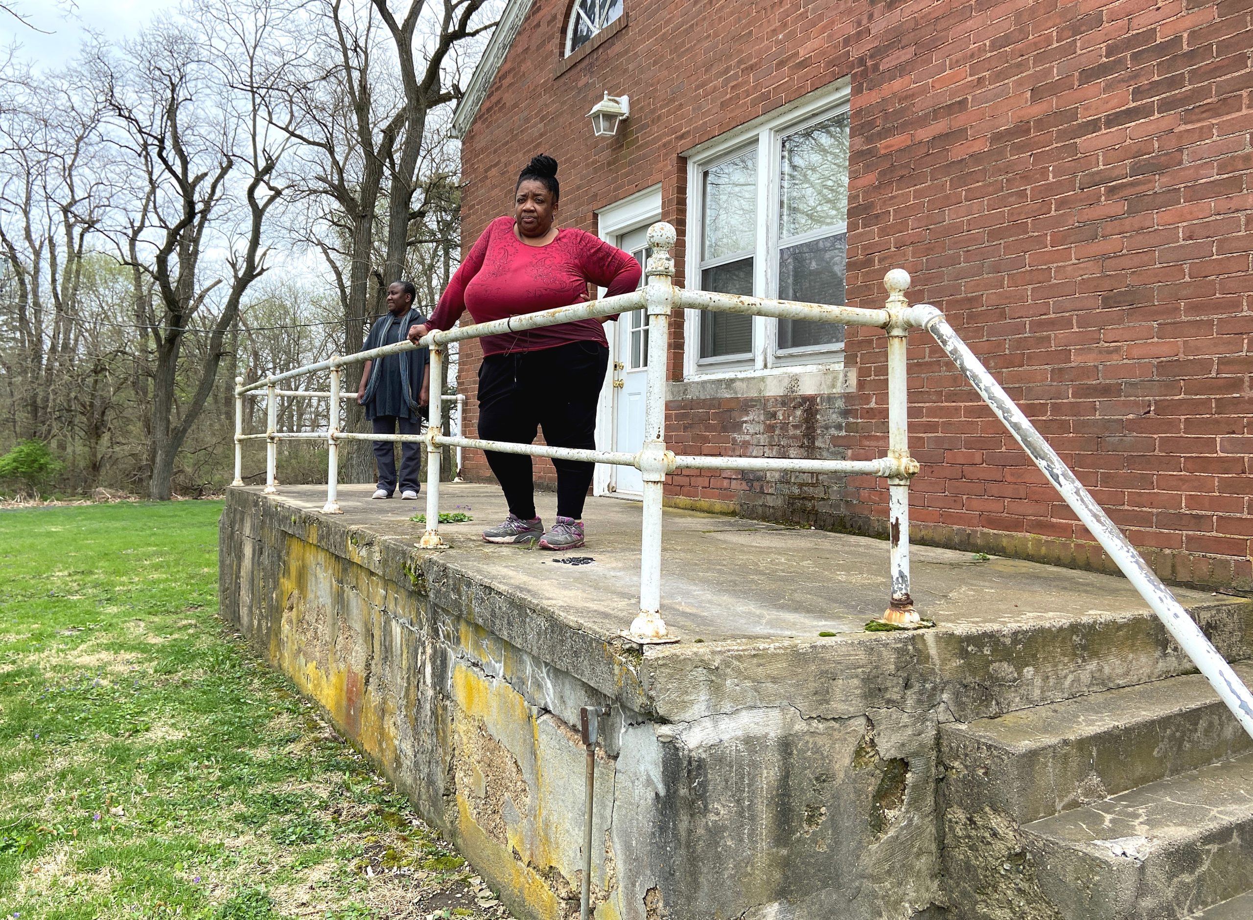Image: Latrelle Bright (left) and Nicole Anderson-Cobb (right) stand on the concrete back patio of the Allerton Park house they occupied during their residency. Bright is Black woman who is wearing a long, gray short-sleeved shirt, gray pants, and a cool-colored striped scarf. She has closely cropped, short hair. She looks off into the distance. Anderson-Cobb is a Black woman who is wearing a red long sleeved shirt, black pants, and a warm colored scarf. Her hair is tied in a top knot on her head. The house is brick with a painted white door and trim. Photo by Jessica Hammie.