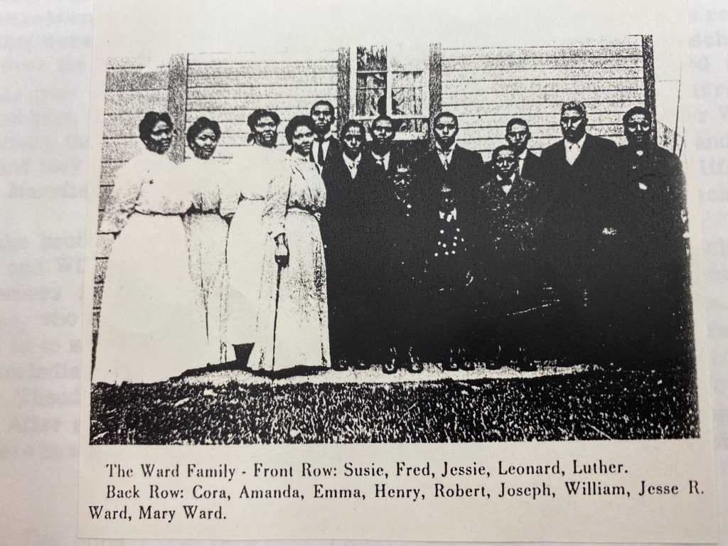 Image: A photo of the Ward family from the Piatt County Archives. A slightly grainy black and white photo reproduction of a Black family standing in front of a wood-sided house with one window centered behind the group. On the left are four Black women, all wearing white dresses with their hair pulled back. On the right, ten Black men and boys wearing dark suits and ties. There is a caption identifying 15 individuals by first name, though the quality of the photo and the poses only show 14 people. Photo courtesy of Nicole Anderson-Cobb. 