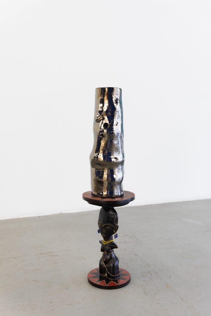 Image: Lola Ogbara, Nikke Vessel, 2021 at The Luminary. A sculpture depicting a wooden Nigerian sculpture of a body that ends at the thighs adorned with jewelry. The figure stands atop and under what appears to be half-inch thick wooden discs. The discs are each painted with a black ten-pointed star with gold highlights and a red background. On the top disc, which is being supported by the figurative sculpture, is a metal sculpture that resembles a thick branch or the base of a thin tree. Image courtesy of Kalaija Mallery.