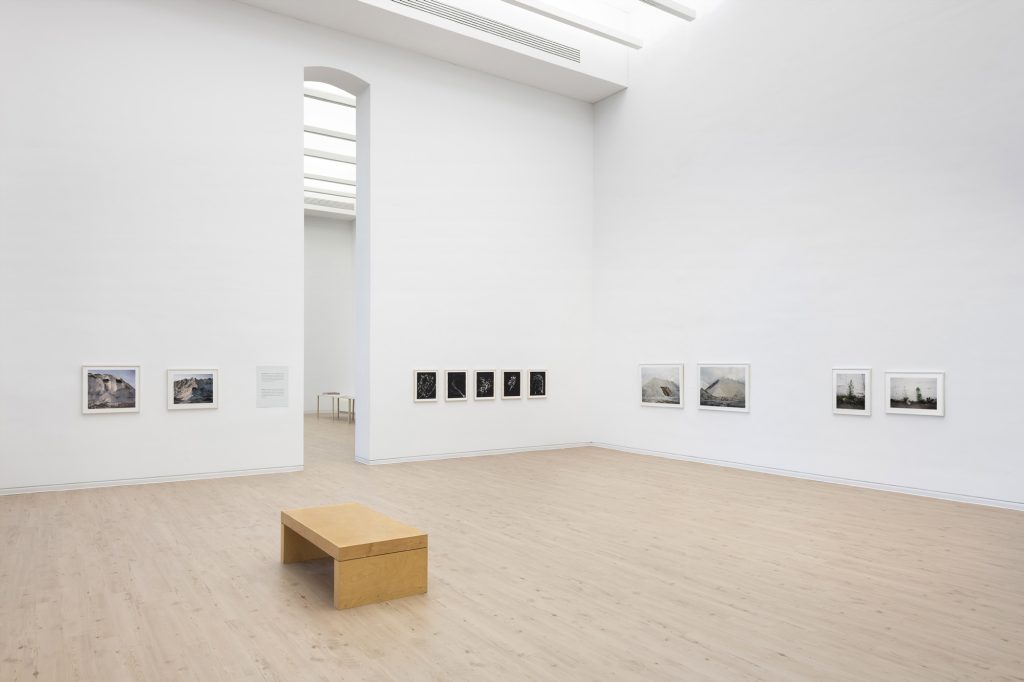 Image: Installation view of 'Views & Scenes'. A large, voluminous gallery space with a tall, narrow doorway leading to the next gallery holds two bodies of work by Jin Lee. A selection of images from 'Weed' and 'Salt Mountain' are mounted on the lower half of tall white walls. Photo by James Prinz, courtesy of the Chicago Cultural Center.